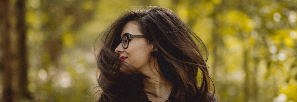 A woman with long, dark, windswept hair and wearing glasses, facing to the side.